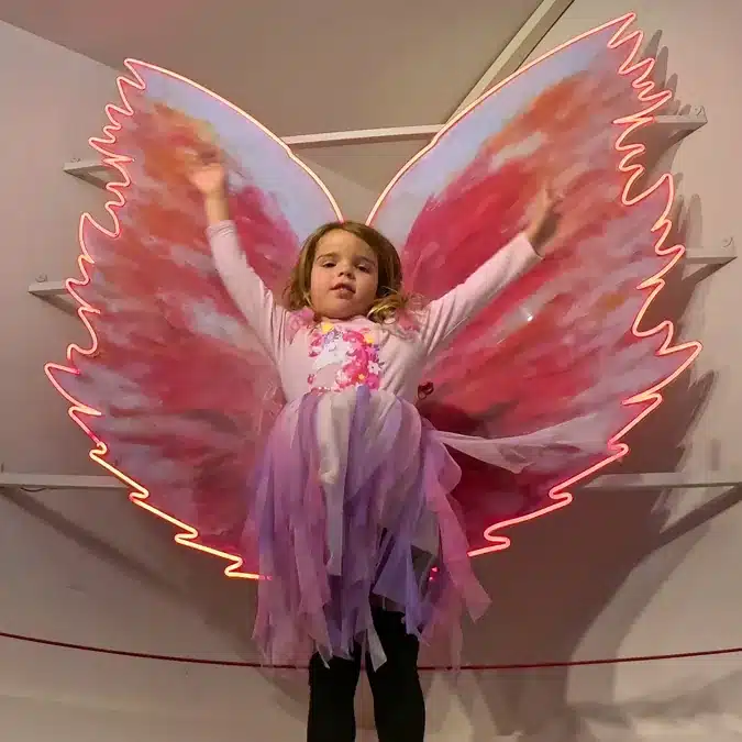 IKONO Madrid: una aventura inmersiva para despertar tu creatividad en familia 3 Niña con alas de hada frente a una instalación de neón en Ikono Madrid con niños